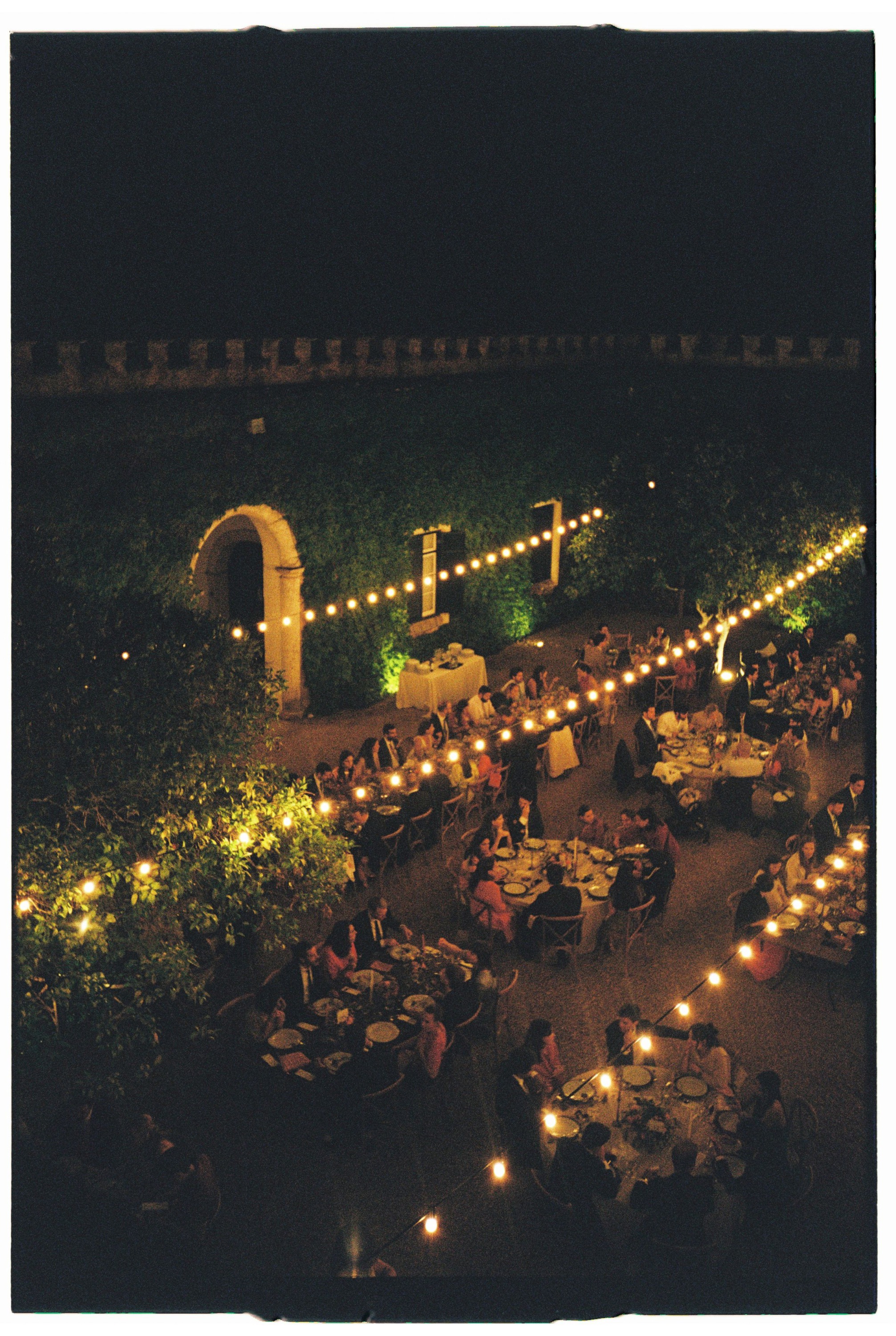 Outdoor evening dinner reception with string lights illuminating tables and guests under a romantic archway.